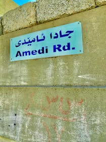 A blue metal sign attached to a concrete wall displays the text 'Amedi Rd.' in both English and Arabic script. The wall has a rough texture and appears weathered, with graffiti in red below the sign.