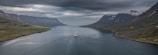 A large cruise ship is sailing along a wide, calm fjord surrounded by steep, shadowy mountains. The sky is overcast, with thick clouds casting a moody atmosphere over the scene. The mountains have patches of snow near their peaks, and the water is a deep, reflective gray that mirrors the overcast sky.