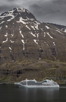A large cruise ship is navigating calm waters with a massive mountain landscape in the background. The mountain is snow-capped at its peak and has patches of snow trailing down its rugged slopes. The sky is overcast with grey clouds.
