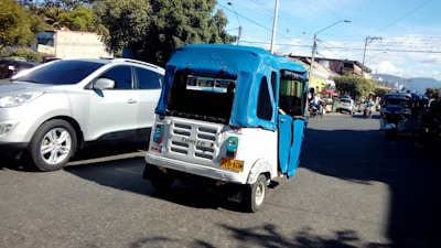 A bustling street scene featuring a blue and white tuk-tuk with a license plate, surrounded by other vehicles including a silver car. Trees and buildings line the street under a blue sky with scattered clouds. Overhead wires and streetlights are visible.
