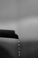 A close-up of a dark, textured pipe with a curved end from which a stream of water is dripping downwards. The background is out of focus, appearing as various shades of gray, giving a soft, blurred effect.