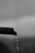 A close-up of a dark, textured pipe with a curved end from which a stream of water is dripping downwards. The background is out of focus, appearing as various shades of gray, giving a soft, blurred effect.