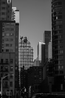 A black and white cityscape featuring a cluster of tall buildings with varying architectural styles. The skyline is densely packed, with sunlight creating shadows and highlights on the structures. The foreground includes a few vehicles and streetlights, adding to the urban atmosphere.
