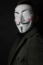 A person wearing a white mask with black, stylized features including a mustache, goatee, and thick eyebrows, reminiscent of a Guy Fawkes mask. The mask has pink blush on the cheeks. The person is dressed in a dark, textured coat, and the background is black, creating a mysterious and enigmatic atmosphere.
