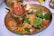A brass plate containing various items used in a traditional ritual. An apple is placed on a small brass pot surrounded by betel leaves, marigold petals, round clay objects, and a sweet dessert typically used in offerings.