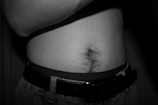A close-up of a person's midsection showing their navel, lower abdomen, and part of their jeans with a black belt. The image is in black and white, emphasizing the hair around the navel area.