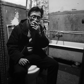A person is sitting on a toilet in a small, dimly lit bathroom. They are wearing glasses and a jacket, and holding a cigarette. The walls appear worn and peeling, with visible plumbing and a bathtub in the background. The atmosphere feels gritty and introspective.
