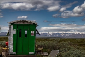 A small green building with white trim stands in an open landscape. The front of the building displays signage with text. It is surrounded by bushes and grass, with distant mountains under a partly cloudy sky in the background.