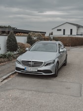A silver luxury car is parked on a residential street. It features modern design elements with sleek lines. In the background, there are well-maintained shrubs and a white, contemporary house with large windows. The sky is overcast, adding a subdued lighting to the scene.