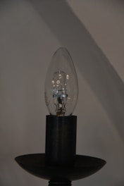 A clear light bulb sits vertically on a dark bulb holder, showcasing the delicate filament and components inside. The background is an out-of-focus wall with beige and gray hues, creating a minimalist and industrial aesthetic.