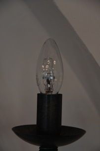 A clear light bulb sits vertically on a dark bulb holder, showcasing the delicate filament and components inside. The background is an out-of-focus wall with beige and gray hues, creating a minimalist and industrial aesthetic.
