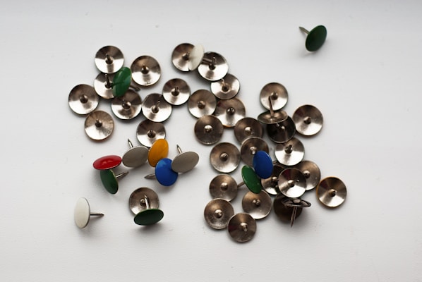 A collection of push pins scattered on a flat surface, with a mix of metallic and colored tops. Some of the push pins are shiny silver, while others have caps in various colors such as green, blue, red, white, and yellow.