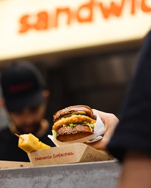 A hand is holding a sandwich with a sesame seed bun filled with what appears to be fried chicken and lettuce. A skewer of fried snacks is visible next to the sandwich. The background is slightly blurred and shows a person wearing a hat, possibly a worker at a food stall or restaurant. The packaging includes visible text with the word 'sandwich'.