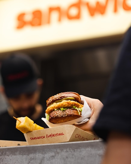 A hand is holding a sandwich with a sesame seed bun filled with what appears to be fried chicken and lettuce. A skewer of fried snacks is visible next to the sandwich. The background is slightly blurred and shows a person wearing a hat, possibly a worker at a food stall or restaurant. The packaging includes visible text with the word 'sandwich'.