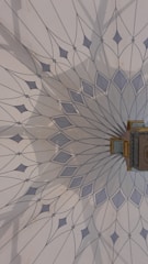 The image features an intricately designed geometric pattern on a wall, resembling a mandala with delicate, pointed shapes radiating from a central point. The color palette includes soft blues and greys, and there is a small, ornate object with gold detailing attached to the wall.