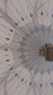 The image features an intricately designed geometric pattern on a wall, resembling a mandala with delicate, pointed shapes radiating from a central point. The color palette includes soft blues and greys, and there is a small, ornate object with gold detailing attached to the wall.