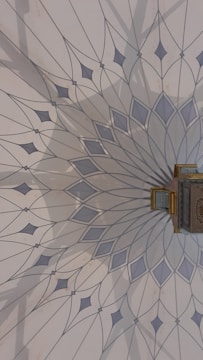 The image features an intricately designed geometric pattern on a wall, resembling a mandala with delicate, pointed shapes radiating from a central point. The color palette includes soft blues and greys, and there is a small, ornate object with gold detailing attached to the wall.