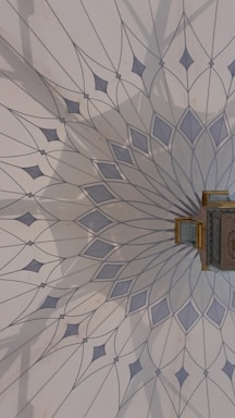 The image features an intricately designed geometric pattern on a wall, resembling a mandala with delicate, pointed shapes radiating from a central point. The color palette includes soft blues and greys, and there is a small, ornate object with gold detailing attached to the wall.