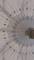 The image features an intricately designed geometric pattern on a wall, resembling a mandala with delicate, pointed shapes radiating from a central point. The color palette includes soft blues and greys, and there is a small, ornate object with gold detailing attached to the wall.