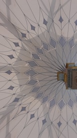 The image features an intricately designed geometric pattern on a wall, resembling a mandala with delicate, pointed shapes radiating from a central point. The color palette includes soft blues and greys, and there is a small, ornate object with gold detailing attached to the wall.