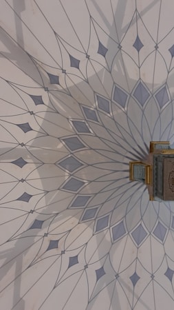The image features an intricately designed geometric pattern on a wall, resembling a mandala with delicate, pointed shapes radiating from a central point. The color palette includes soft blues and greys, and there is a small, ornate object with gold detailing attached to the wall.