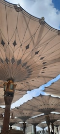Large, ornate canopies with intricate patterns providing shade, designed with a geometric flower-like motif. They are supported by columns, with a blue sky and a few clouds visible above.