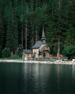 A small, picturesque stone chapel is nestled among tall, dense evergreen trees by the edge of a serene lake. The reflection of the chapel and trees is visible on the calm water surface. The chapel features a steep, pointed roof with a small cross at the top, adding to its quaint and peaceful ambiance. The surrounding area is lush and green, enhancing the tranquil setting.