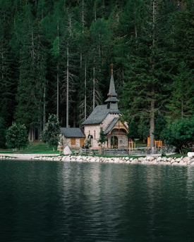 A small, picturesque stone chapel is nestled among tall, dense evergreen trees by the edge of a serene lake. The reflection of the chapel and trees is visible on the calm water surface. The chapel features a steep, pointed roof with a small cross at the top, adding to its quaint and peaceful ambiance. The surrounding area is lush and green, enhancing the tranquil setting.