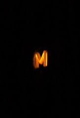 A glowing yellow letter 'M' against a dark background, illuminating the area around it.