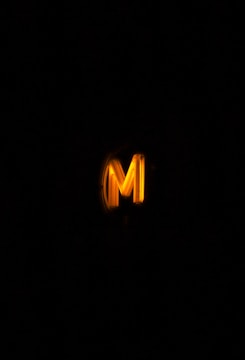 A glowing yellow letter 'M' against a dark background, illuminating the area around it.