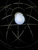 A single white light bulb is encased within a metal wire frame, resembling an atom-like structure with intersecting circles against a dark background.