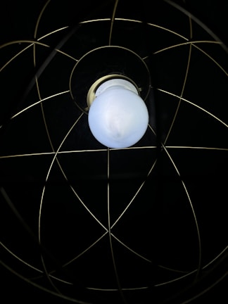 A single white light bulb is encased within a metal wire frame, resembling an atom-like structure with intersecting circles against a dark background.
