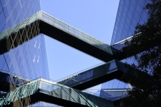 Modern office buildings with reflective glass facades are connected by sleek, horizontal skybridges. The structures create an interplay of lines and reflections against a clear blue sky.