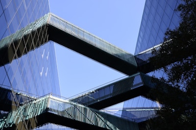 Modern office buildings with reflective glass facades are connected by sleek, horizontal skybridges. The structures create an interplay of lines and reflections against a clear blue sky.