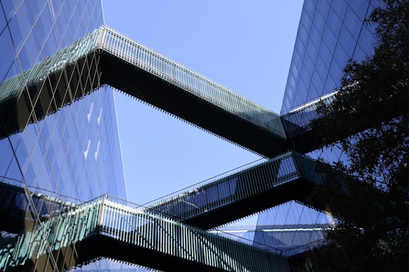 Modern office buildings with reflective glass facades are connected by sleek, horizontal skybridges. The structures create an interplay of lines and reflections against a clear blue sky.