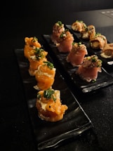 Two black plates are arranged on a dark surface. Each plate holds neatly arranged pieces of sushi rolls garnished with green onions and a creamy sauce. The sushi rolls appear to be wrapped in smoked salmon and a cured meat, possibly prosciutto.