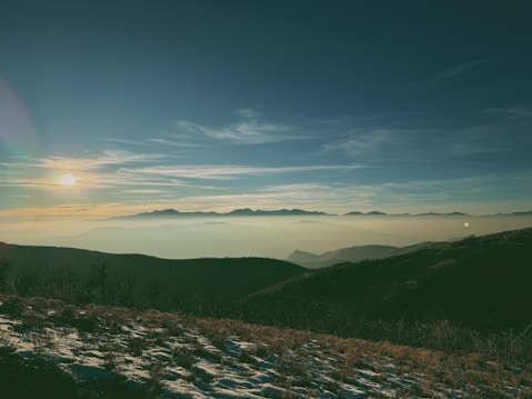 The image displays a serene mountain landscape at sunrise. The sun is low on the horizon, casting a soft glow over distant mountain ranges covered in a light mist. The foreground features snow-dusted, grassy hills with some barren trees, creating a tranquil and expansive view.