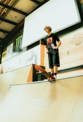 A young person stands on the edge of a skateboard ramp, holding a skateboard. The surroundings include various pieces of art and brand logos on the walls. The ramp and the person create a bold silhouette against a soft, ambient light.