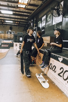 Inside a skate park, three young people are gathered. Two are sitting on a platform with skateboards nearby, while one stands holding a skateboard. The setting includes ramps, advertisements, and a light industrial aesthetic.