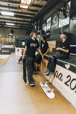 Inside a skate park, three young people are gathered. Two are sitting on a platform with skateboards nearby, while one stands holding a skateboard. The setting includes ramps, advertisements, and a light industrial aesthetic.