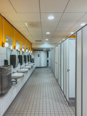 A spacious public restroom with a row of modern white sinks and mirrors on the left and a series of closed stall doors on the right. The floor is covered with gray tiles and the ceiling has recessed lighting. Bright yellow walls add color to the space, and there are stainless steel hand dryers and waste bins near the sinks.