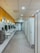 A spacious public restroom with a row of modern white sinks and mirrors on the left and a series of closed stall doors on the right. The floor is covered with gray tiles and the ceiling has recessed lighting. Bright yellow walls add color to the space, and there are stainless steel hand dryers and waste bins near the sinks.