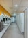A spacious public restroom with a row of modern white sinks and mirrors on the left and a series of closed stall doors on the right. The floor is covered with gray tiles and the ceiling has recessed lighting. Bright yellow walls add color to the space, and there are stainless steel hand dryers and waste bins near the sinks.