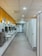 A spacious public restroom with a row of modern white sinks and mirrors on the left and a series of closed stall doors on the right. The floor is covered with gray tiles and the ceiling has recessed lighting. Bright yellow walls add color to the space, and there are stainless steel hand dryers and waste bins near the sinks.