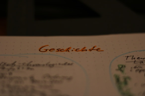 A close-up view of a page with handwritten notes. The word 'Geschichte' is prominently written in orange ink at the top of the page, surrounded by dotted paper. Other text and diagrams are visible but blurred in the background.