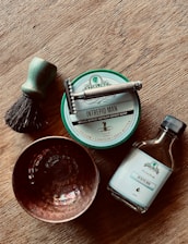A wooden surface holds a collection of shaving items, including a brush with a green handle and dark bristles, a silver safety razor, a tin of 'Intrepid Man' shave soap, a bottle of 'Intrepid Man' aftershave splash, and a textured copper bowl. The arrangement suggests a classic, traditional grooming setup.