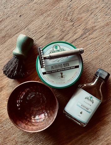 A wooden surface holds a collection of shaving items, including a brush with a green handle and dark bristles, a silver safety razor, a tin of 'Intrepid Man' shave soap, a bottle of 'Intrepid Man' aftershave splash, and a textured copper bowl. The arrangement suggests a classic, traditional grooming setup.