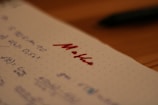 Notebook paper with mathematical equations written in blue ink, alongside the word 'Maths' written prominently in red ink. A pen, placed out of focus, lies on a wooden table in the background.