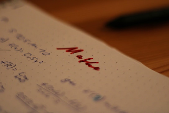 Notebook paper with mathematical equations written in blue ink, alongside the word 'Maths' written prominently in red ink. A pen, placed out of focus, lies on a wooden table in the background.