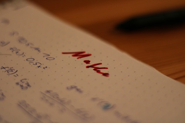 Notebook paper with mathematical equations written in blue ink, alongside the word 'Maths' written prominently in red ink. A pen, placed out of focus, lies on a wooden table in the background.